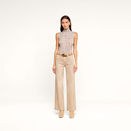 A woman with straight brown hair and a neutral expression stands against a white background, wearing the Ceci Sleeveless Turtleneck in a grey leopard print, paired with beige wide-leg pants featuring a belt and matching heeled boots.