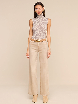 A woman stands against a beige backdrop, wearing the Ceci Sleeveless Turtleneck—a grey leopard print top—paired with wide-leg beige pants, a tan belt, and beige boots. She accessorizes with gold earrings and has long dark hair.