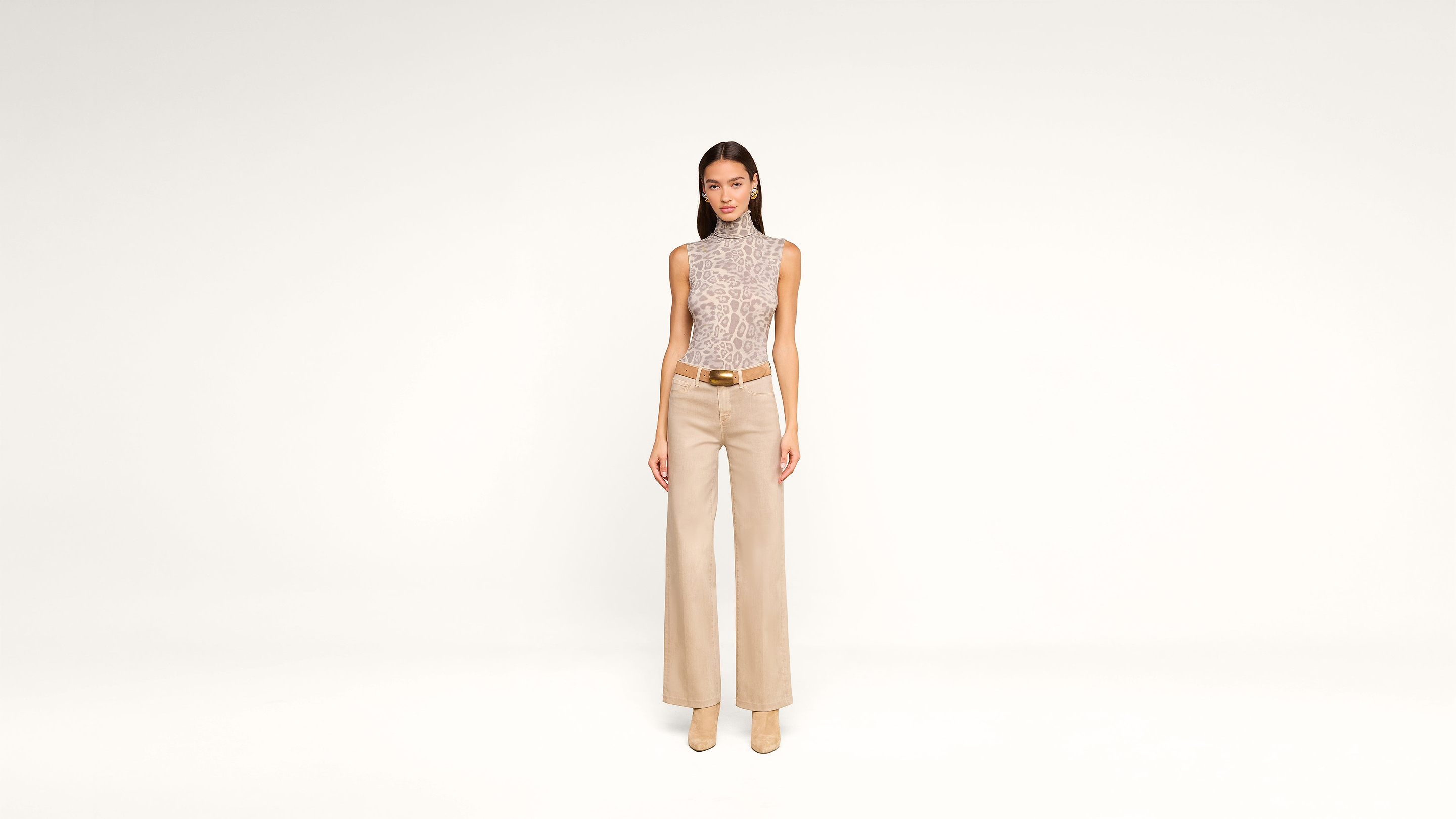 A woman stands facing forward against a plain white background, wearing a sleeveless, patterned turtleneck top, wide-leg beige trousers, a brown belt, and beige heeled shoes.