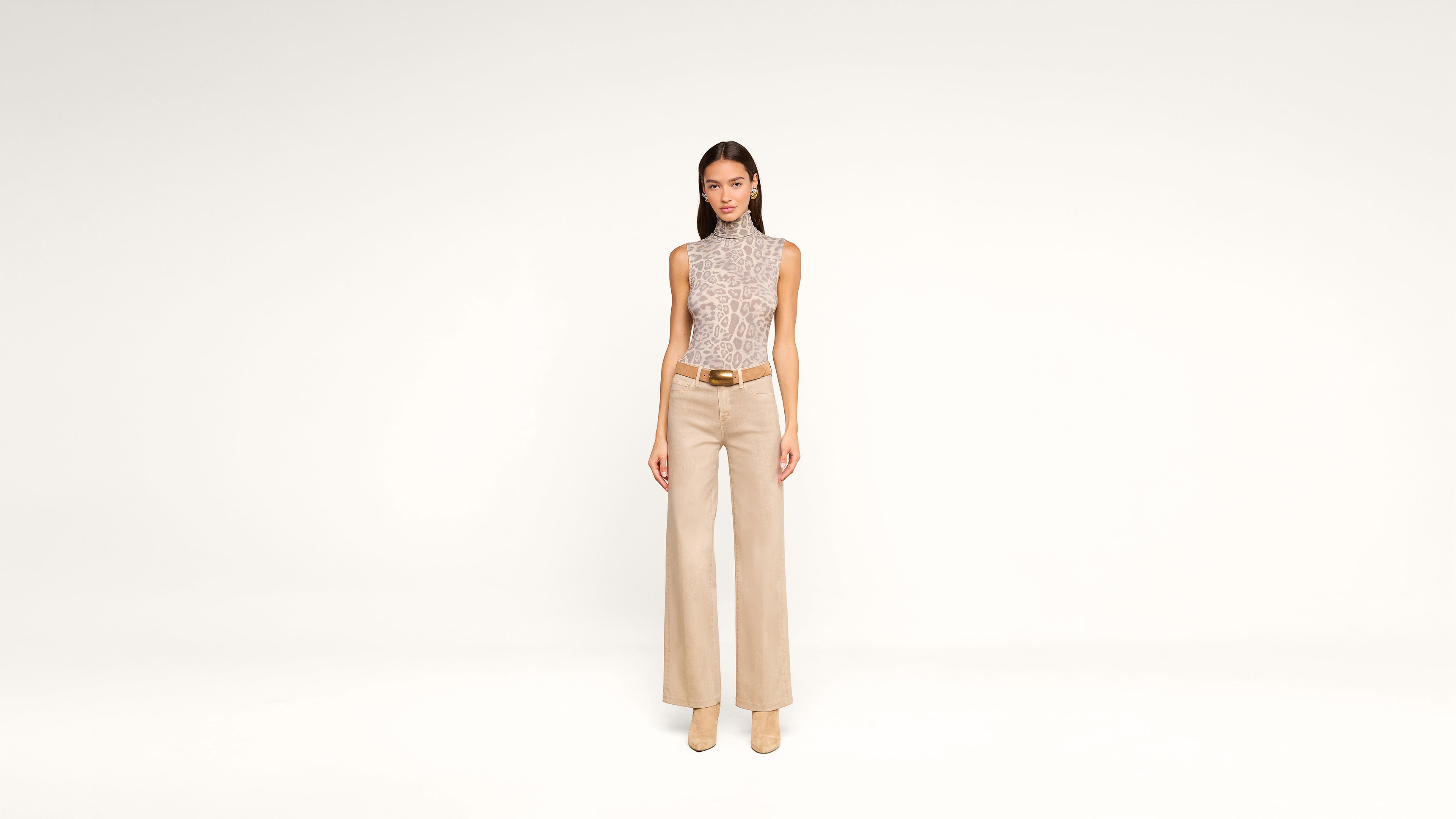 A woman stands facing forward against a plain white background, wearing a sleeveless, patterned turtleneck top, wide-leg beige trousers, a brown belt, and beige heeled shoes.