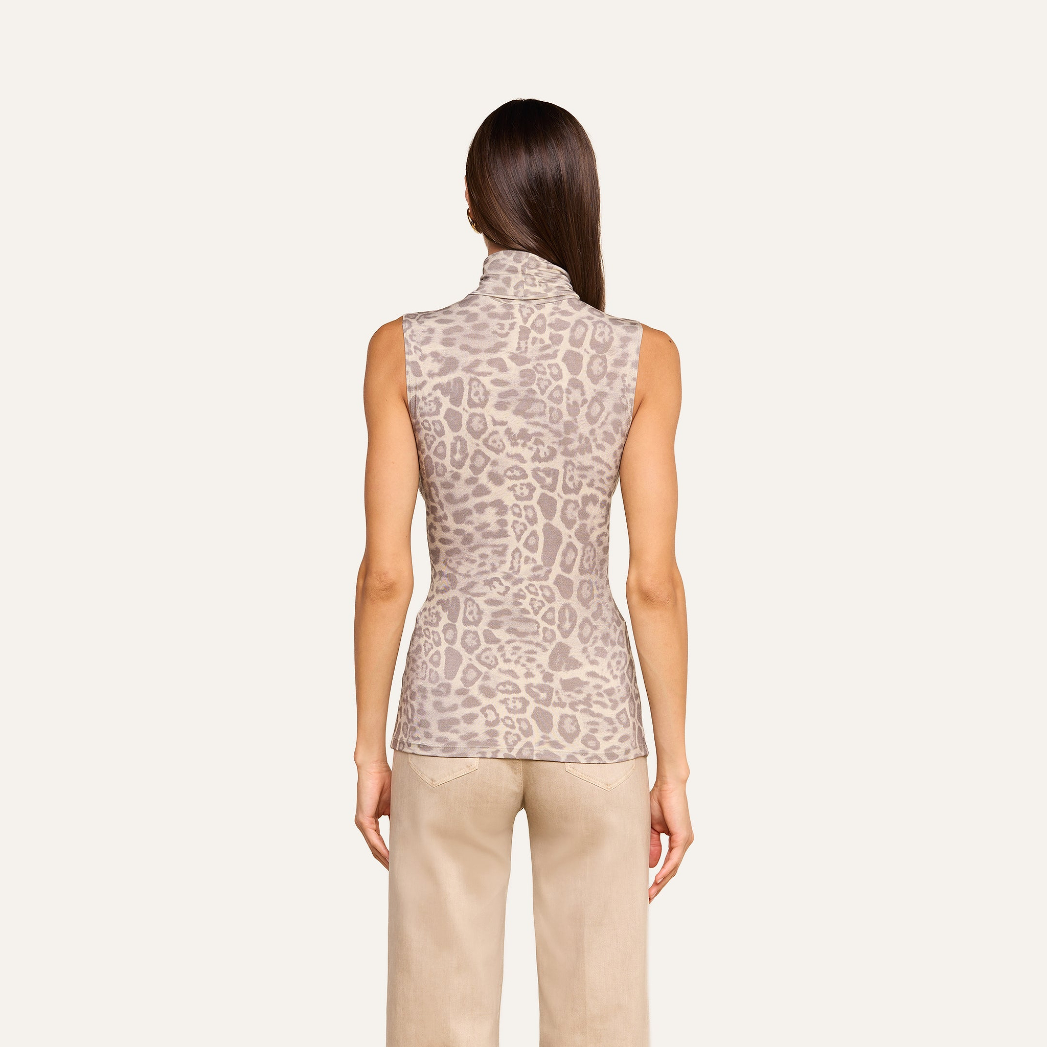 A woman stands with her back to the camera, wearing the Ceci Sleeveless Turtleneck in a light brown animal print and beige pants, set against a plain light background.