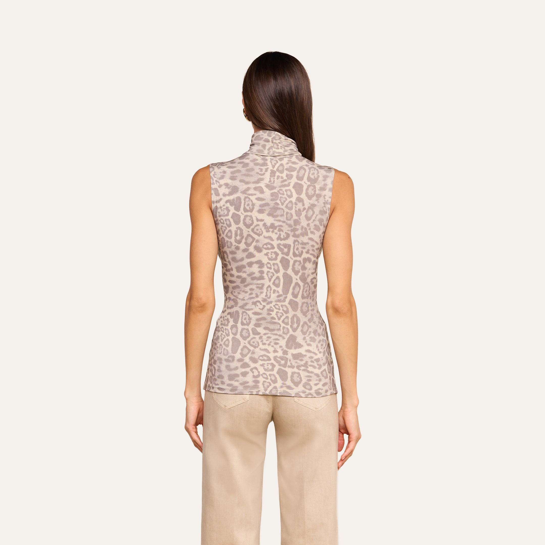 A woman stands with her back to the camera, wearing the Ceci Sleeveless Turtleneck in a light brown animal print and beige pants, set against a plain light background.