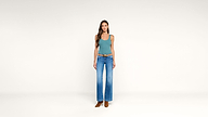 Woman stands against a plain white background, wearing a teal tank top, blue high-waisted wide-leg jeans, a brown belt, and brown boots. Her hair is down and she is looking forward with hands at her sides.