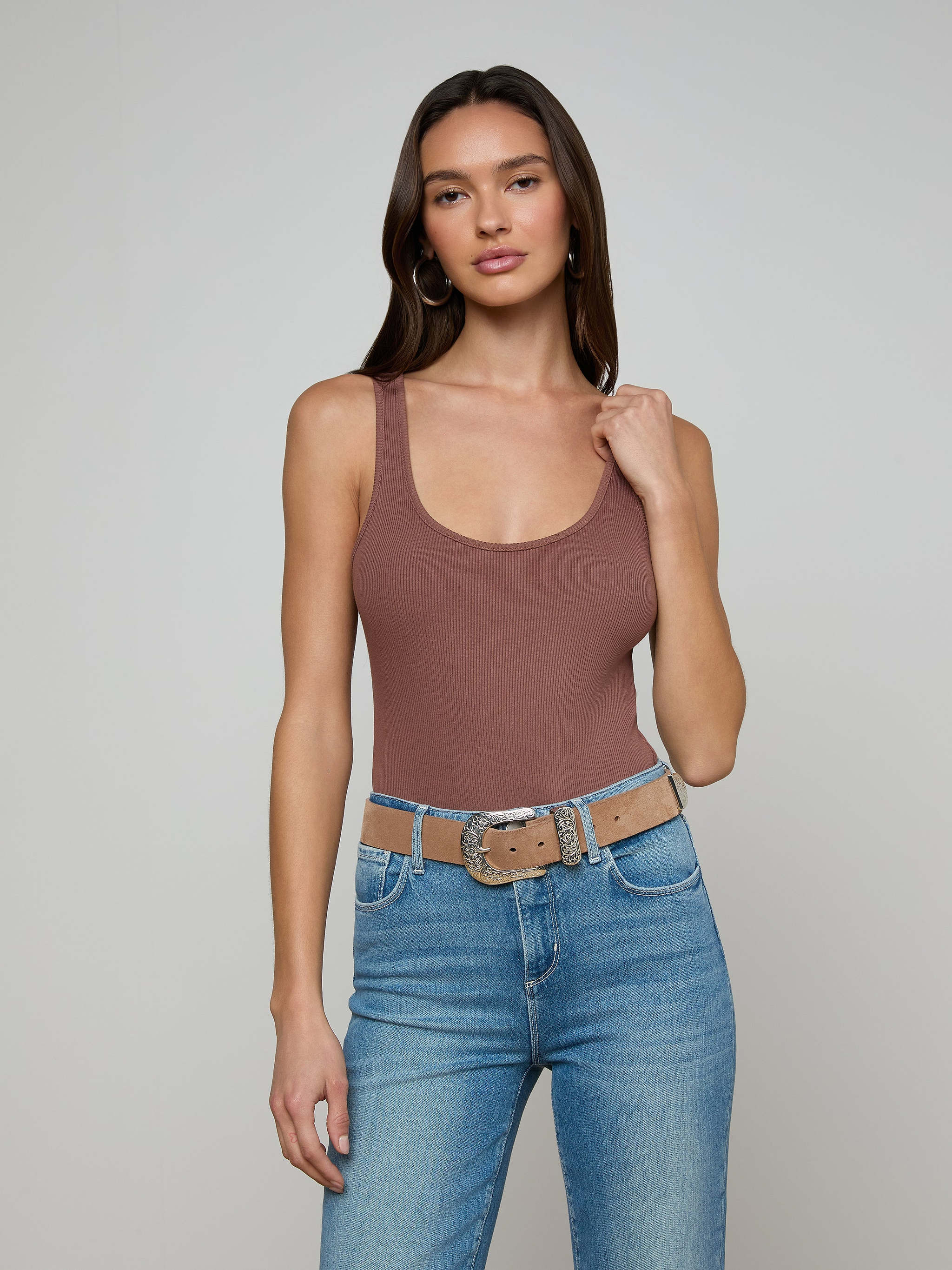 Woman with long brown hair wearing the Nica Scoopneck Bodysuit in brown, blue jeans, and a beige belt with a silver buckle, standing against a plain light gray background.