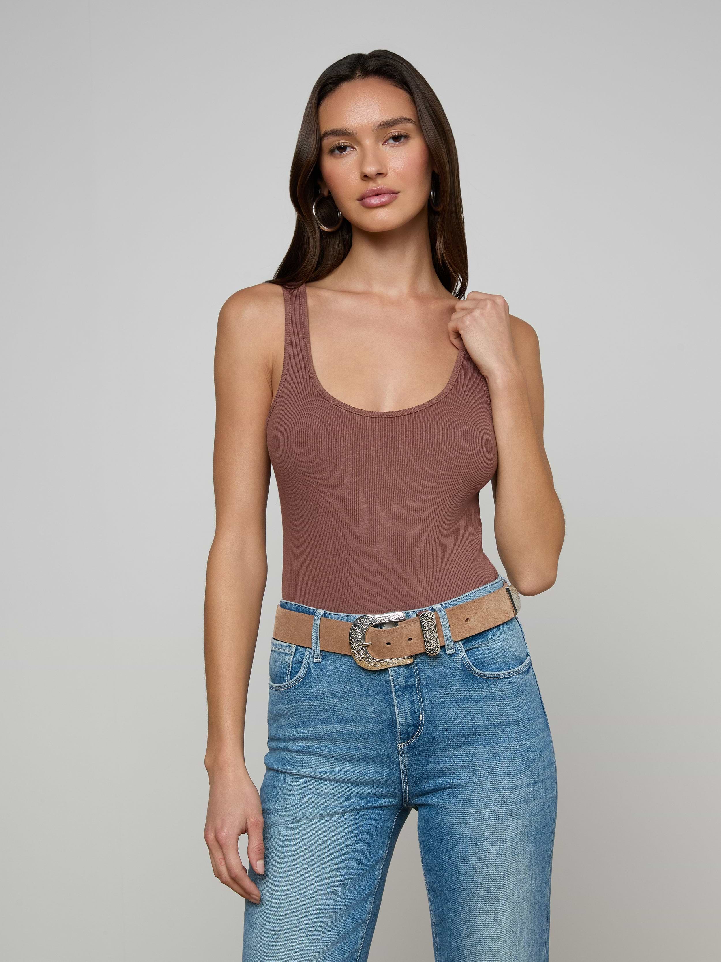Woman with long brown hair wearing the Nica Scoopneck Bodysuit in brown, blue jeans, and a beige belt with a silver buckle, standing against a plain light gray background.