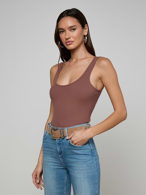 A woman with long brown hair models the Nica Scoopneck Bodysuit and high-waisted blue jeans with a beige belt, standing against a plain light gray background.