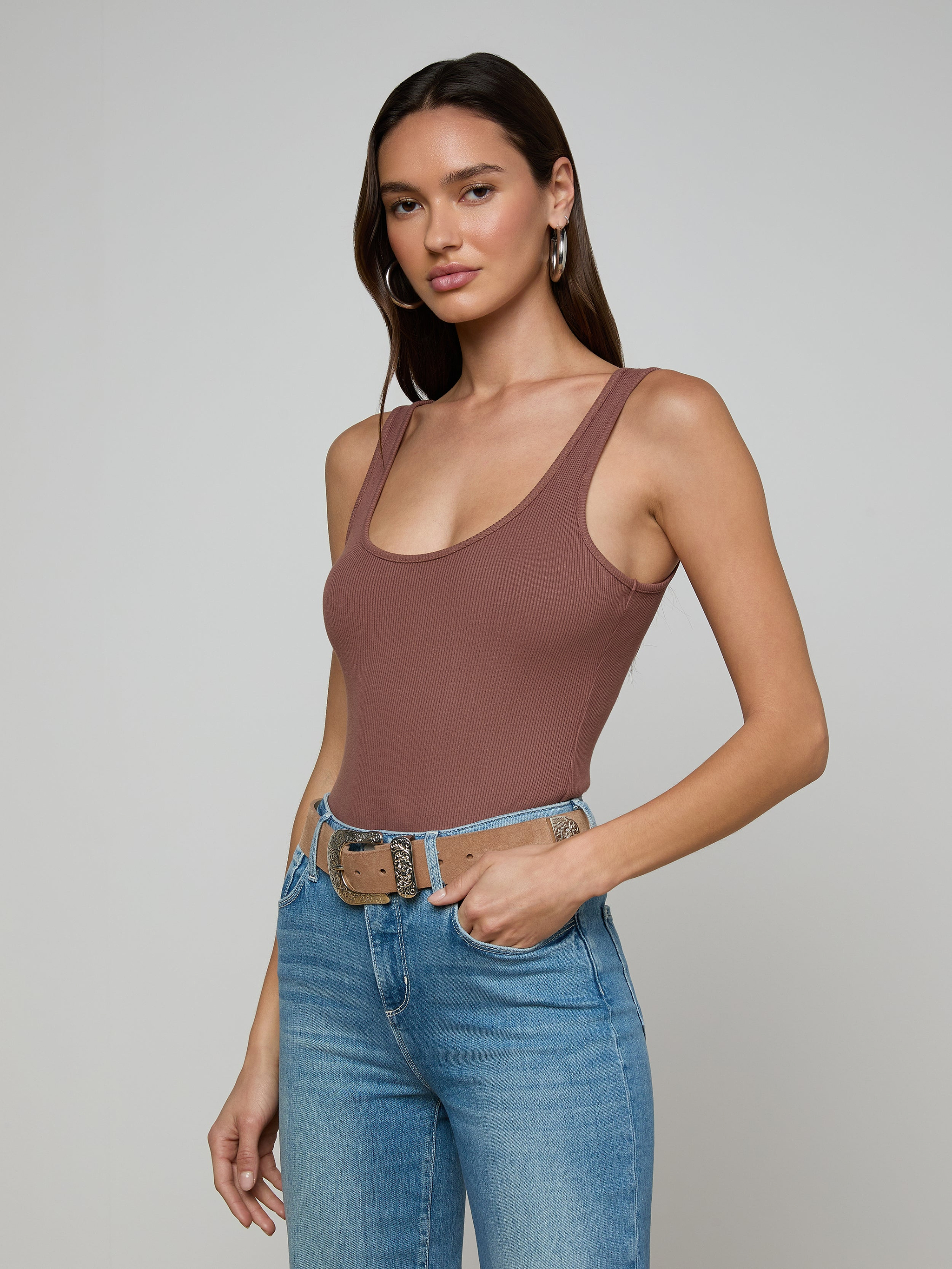 A woman with long brown hair models the Nica Scoopneck Bodysuit and high-waisted blue jeans with a beige belt, standing against a plain light gray background.
