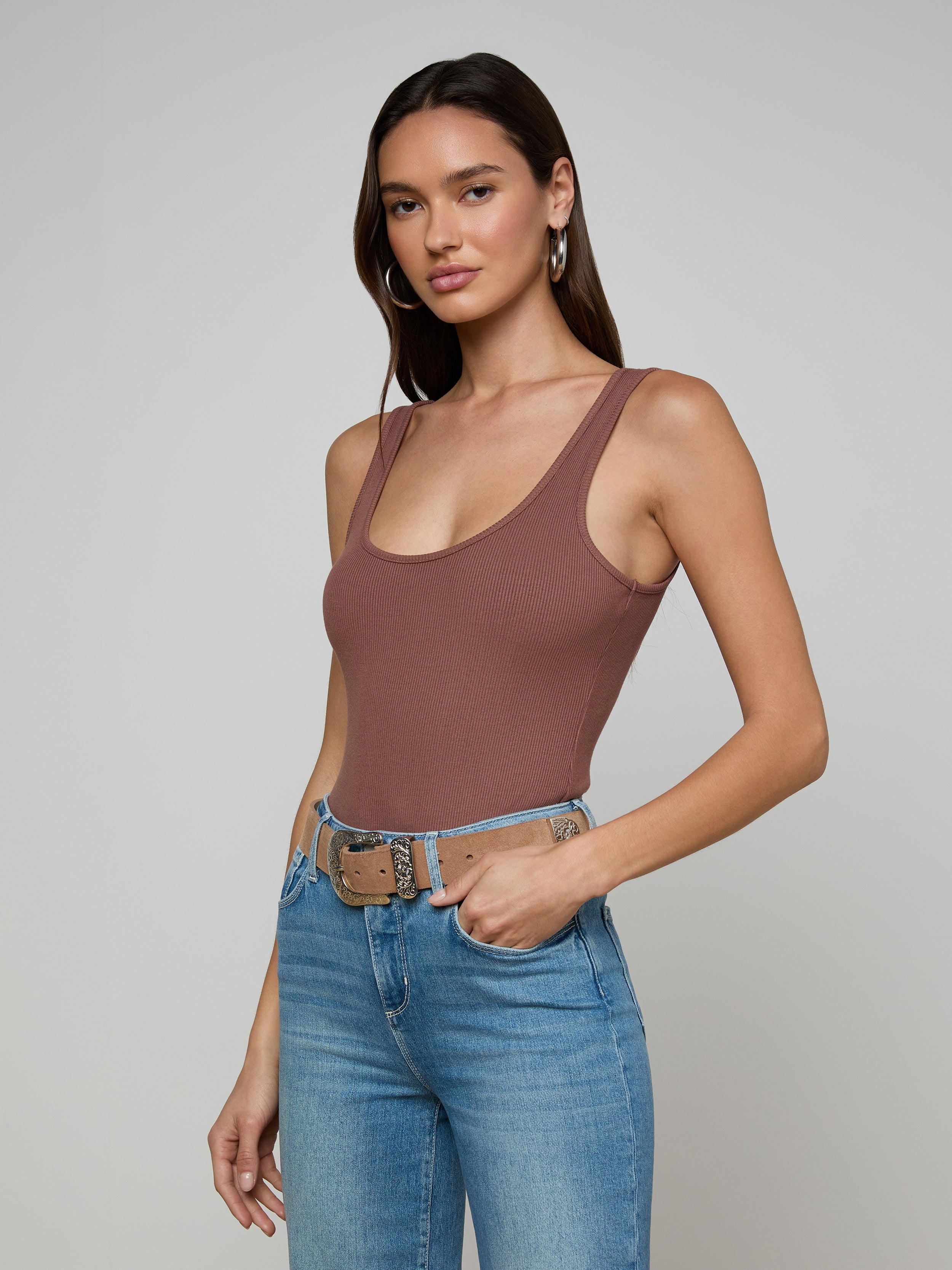 A woman with long brown hair models the Nica Scoopneck Bodysuit and high-waisted blue jeans with a beige belt, standing against a plain light gray background.