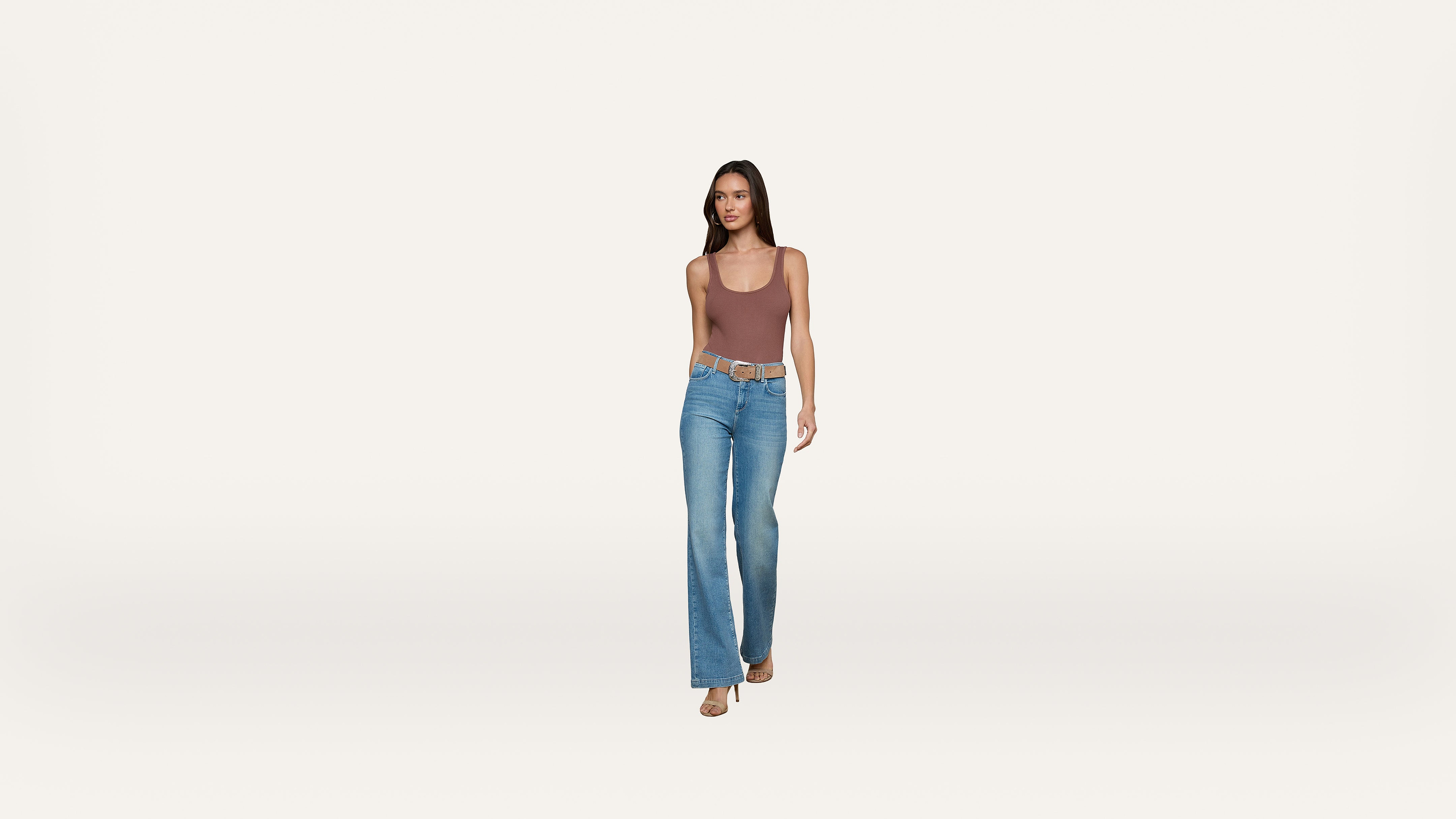 A woman wearing a brown tank top, blue flared jeans, and sandals walks forward against a plain white background.