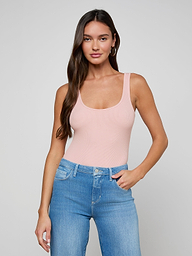 A woman with long brown hair stands against a plain off-white background, looking at the camera with a neutral expression while wearing the Nica Scoopneck Bodysuit in light pink and blue jeans.