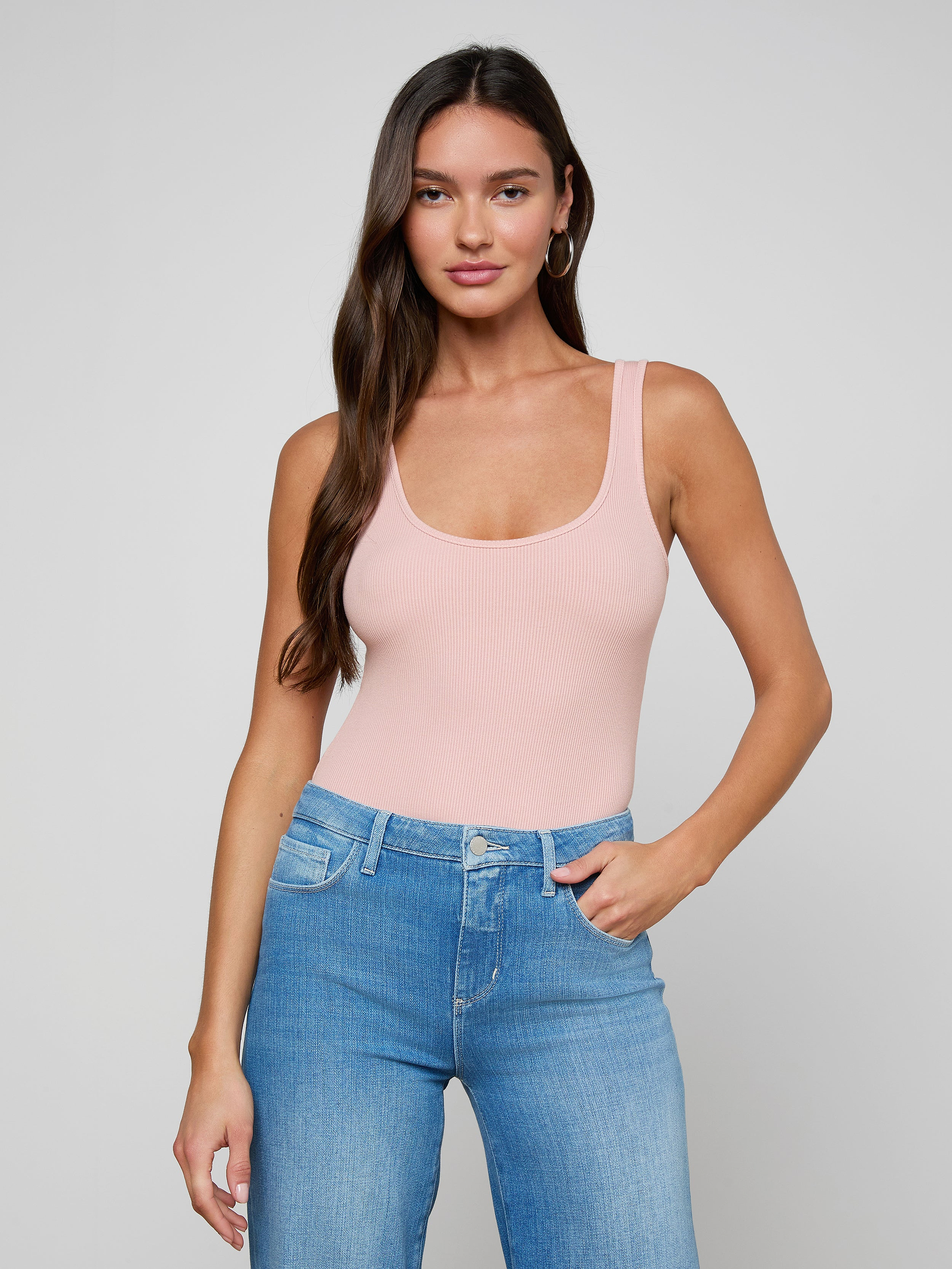 A woman with long brown hair stands against a plain off-white background, looking at the camera with a neutral expression while wearing the Nica Scoopneck Bodysuit in light pink and blue jeans.