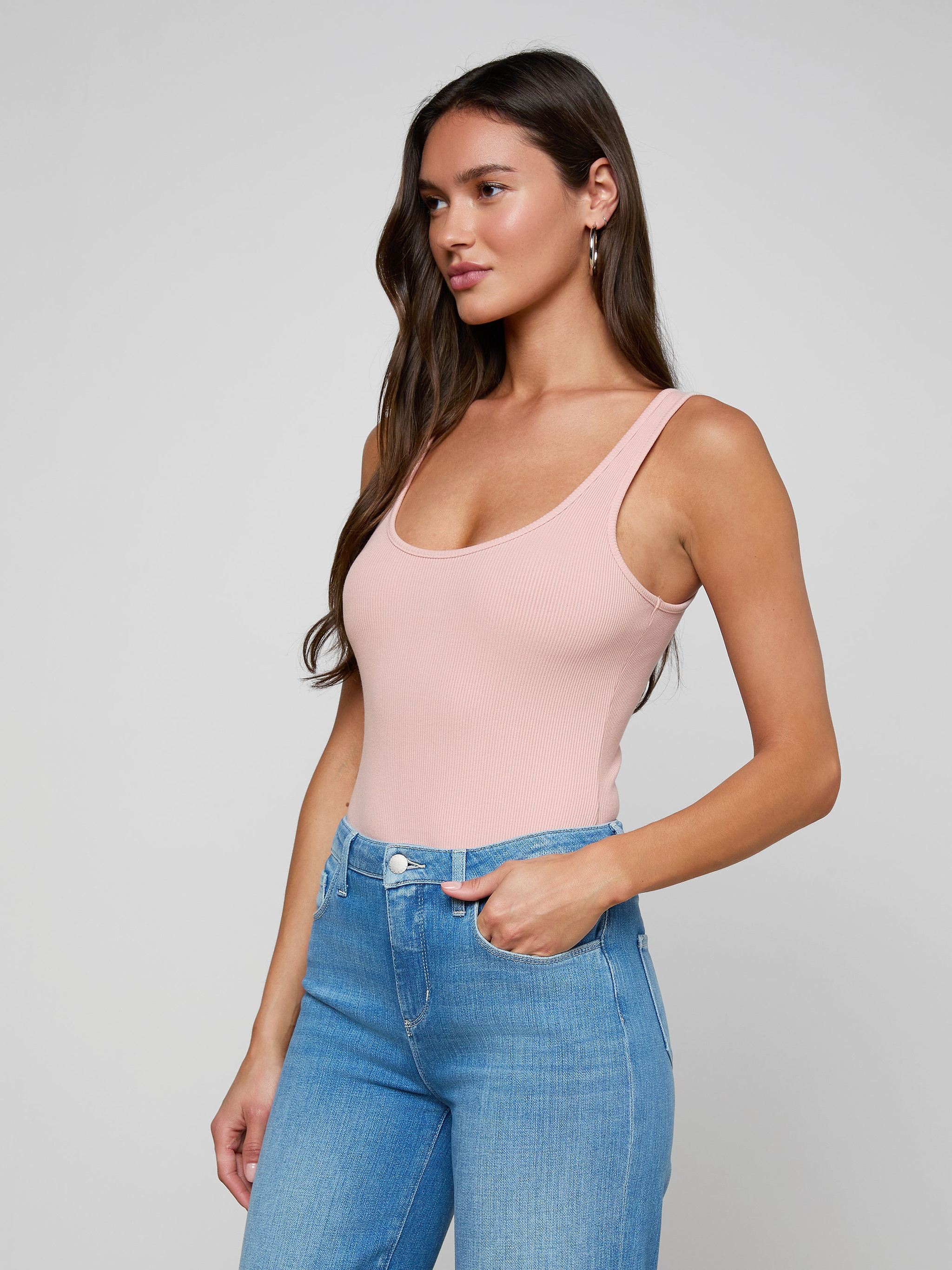 A woman with long brown hair wears the Nica Scoopneck Bodysuit in light pink paired with blue jeans, standing with one hand in her pocket and gazing to the side against a plain light background.