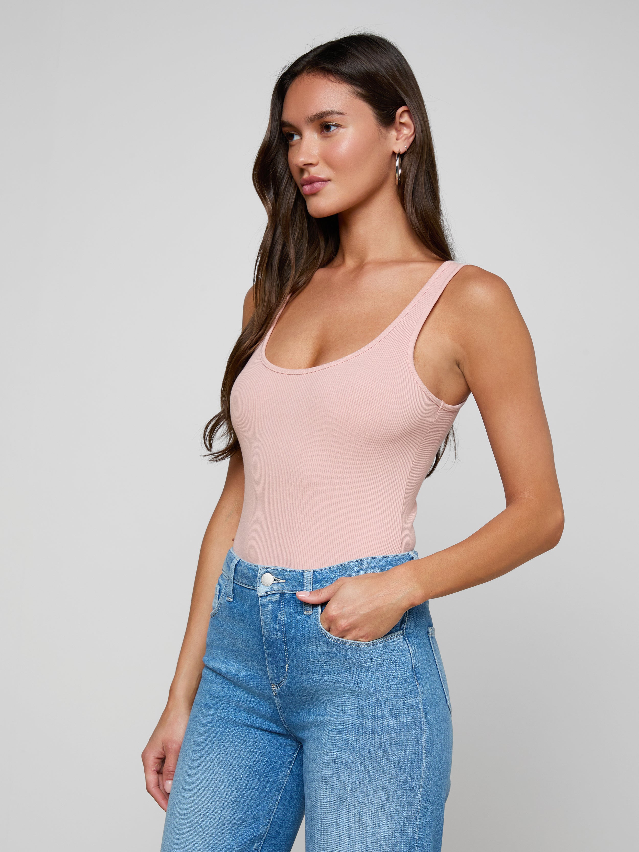 A woman with long brown hair wears the Nica Scoopneck Bodysuit in light pink paired with blue jeans, standing with one hand in her pocket and gazing to the side against a plain light background.