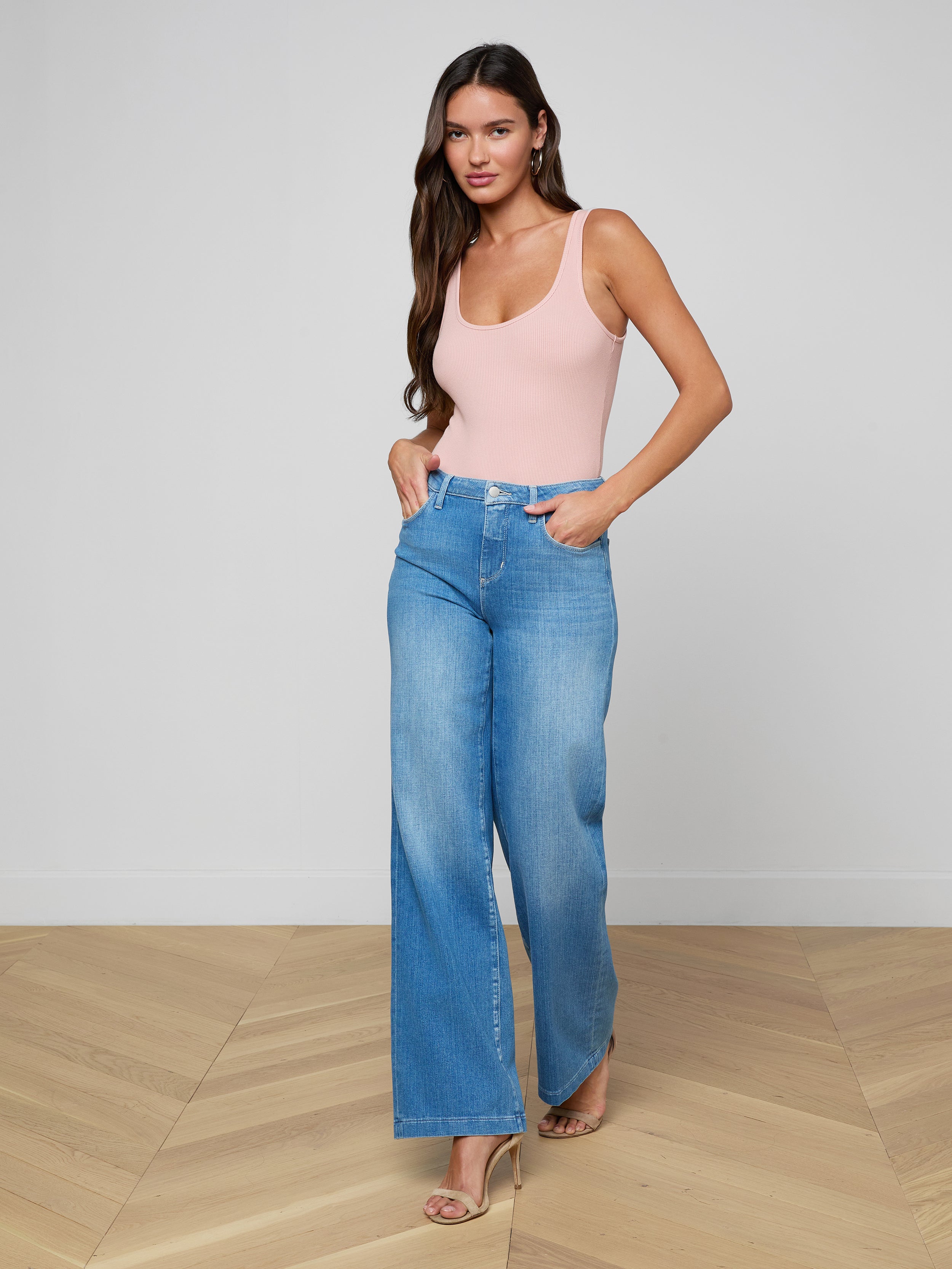 A woman wearing the Nica Scoopneck Bodysuit in light pink, paired with high-waisted blue wide-leg jeans and beige heels, stands against a plain white background, looking at the camera with her hands in her pockets.