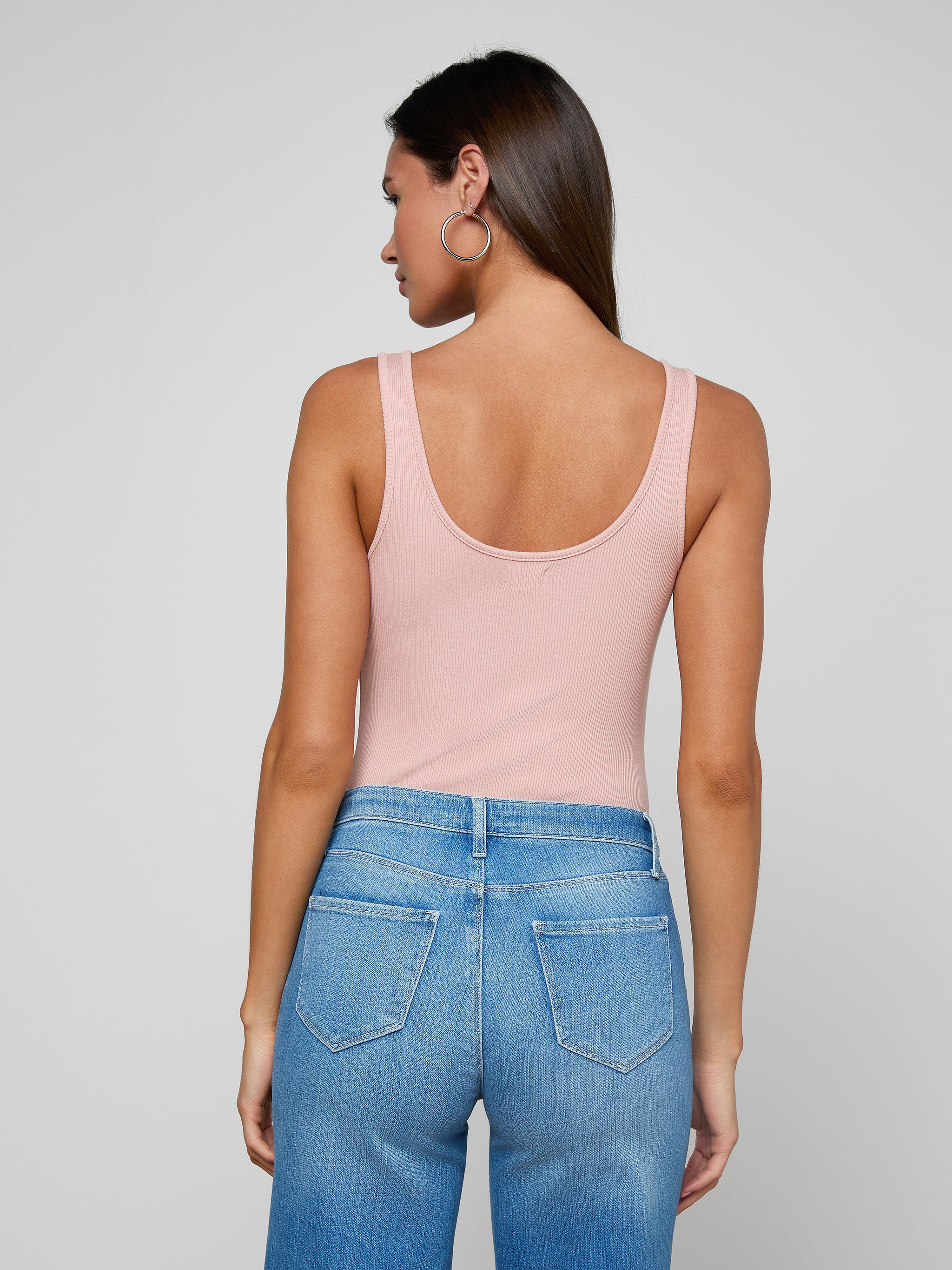 Woman with long brown hair and large hoop earrings, wearing the Nica Scoopneck Bodysuit in light pink high stretch rib and blue high-waisted jeans, shown from the back against an off-white background.