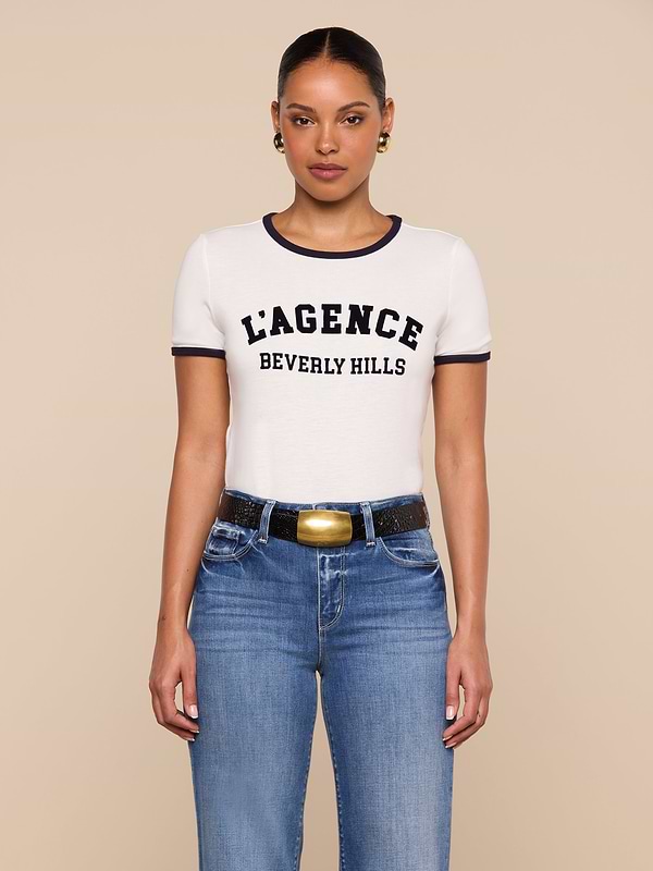 A woman stands against a beige background wearing the relaxed fit white Tommie Crewneck Tee with black trim that reads L’AGENCE BEVERLY HILLS, paired with blue jeans, a black belt with a gold buckle, and gold hoop earrings.