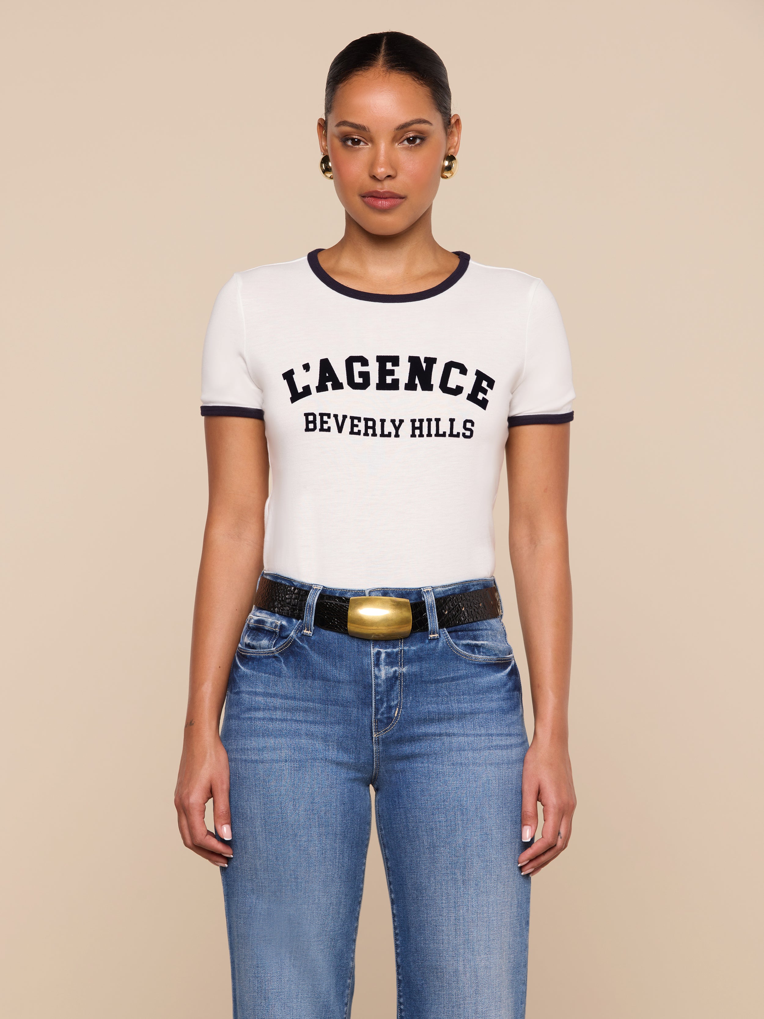 A woman stands against a beige background wearing the relaxed fit white Tommie Crewneck Tee with black trim that reads L’AGENCE BEVERLY HILLS, paired with blue jeans, a black belt with a gold buckle, and gold hoop earrings.