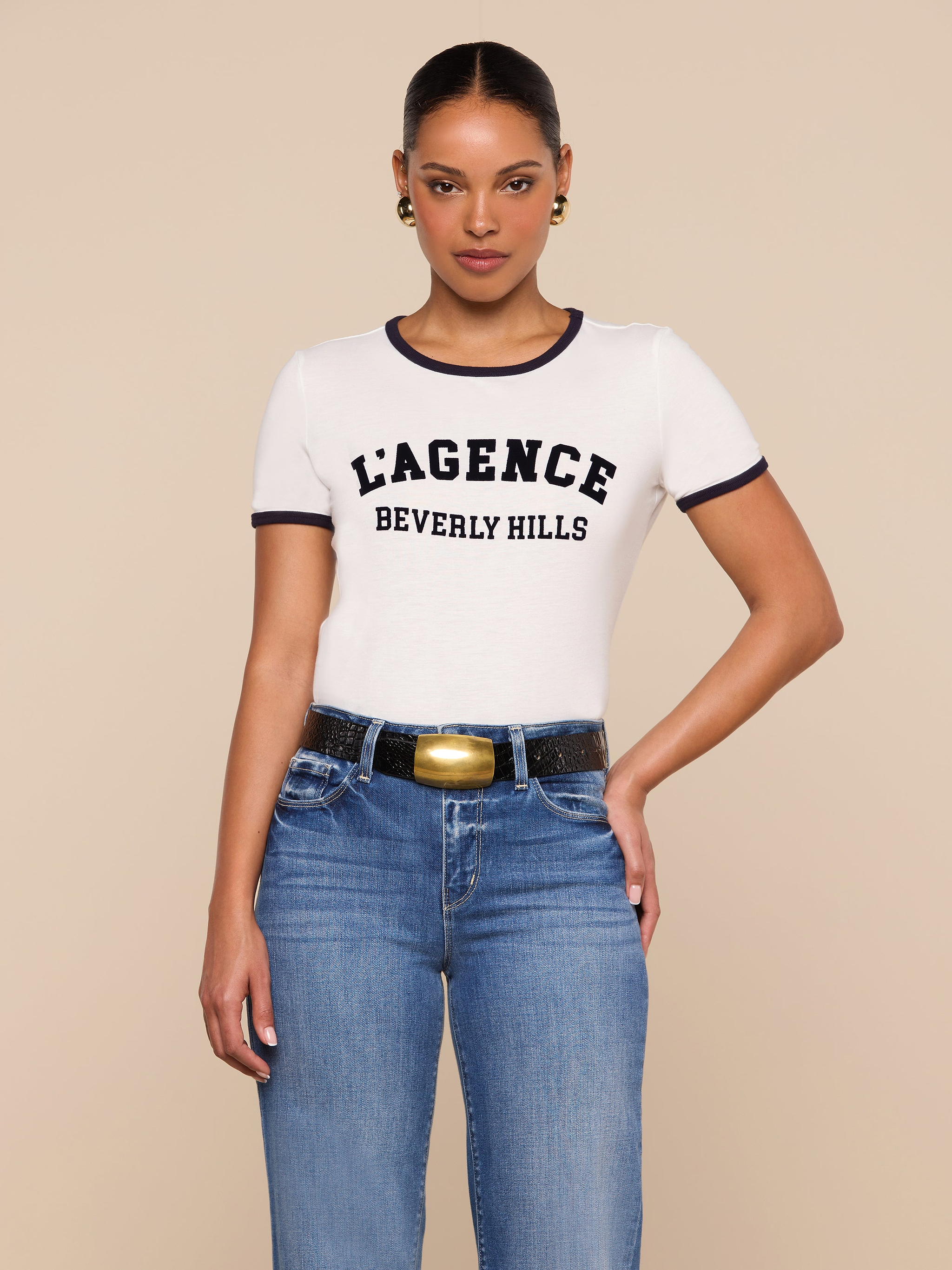 A woman stands against a beige background in a relaxed fit white Tommie Crewneck Tee with LAGENCE BEVERLY HILLS in bold black letters, blue jeans, a black belt with a gold buckle, and gold earrings, looking confidently at the camera.