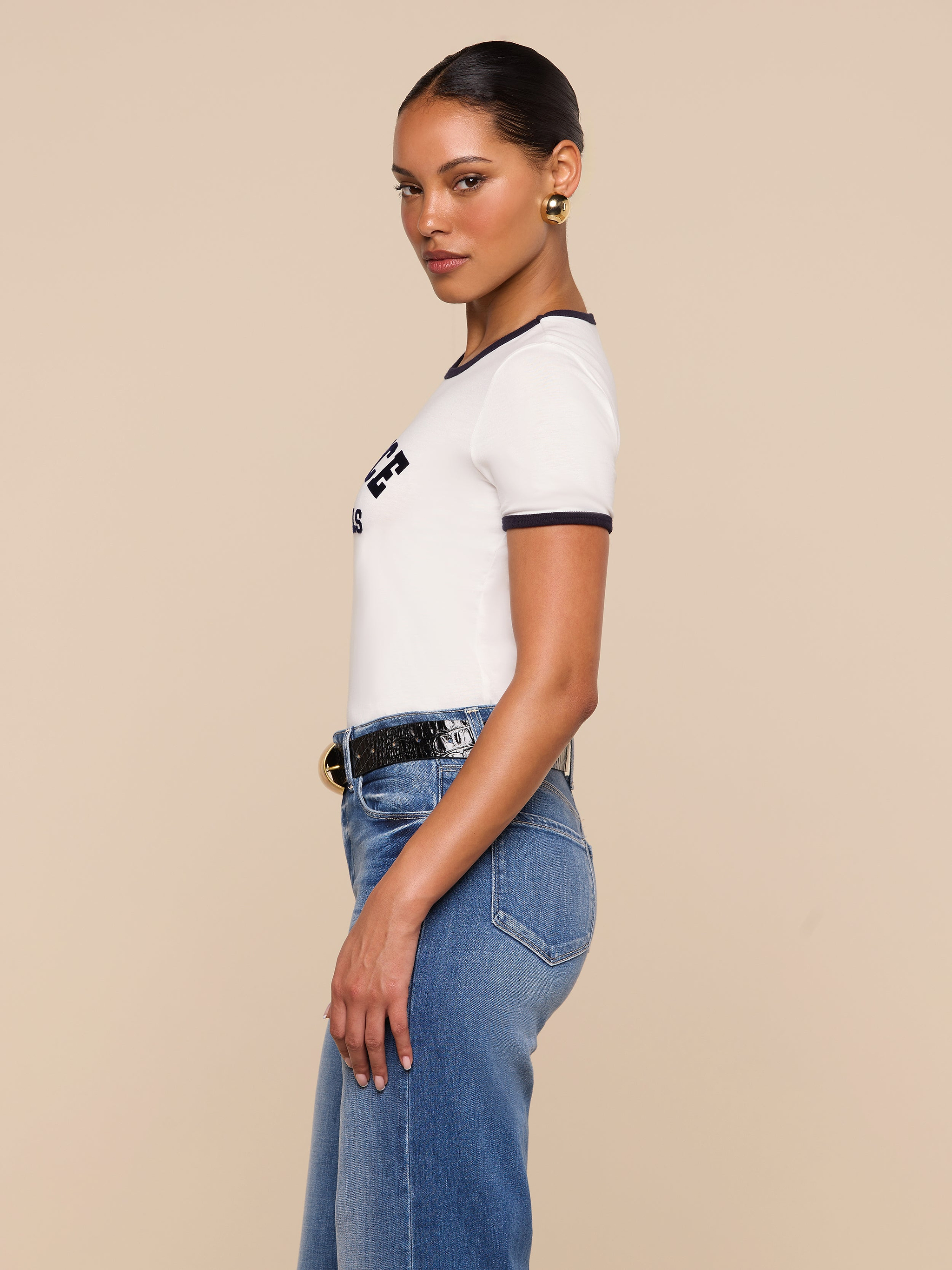 A woman with slicked-back hair poses in profile against a beige background, wearing the Tommie Crewneck Tee—a relaxed fit white Supima cotton t-shirt with black trim—paired with blue jeans, a black belt, and hoop earrings.
