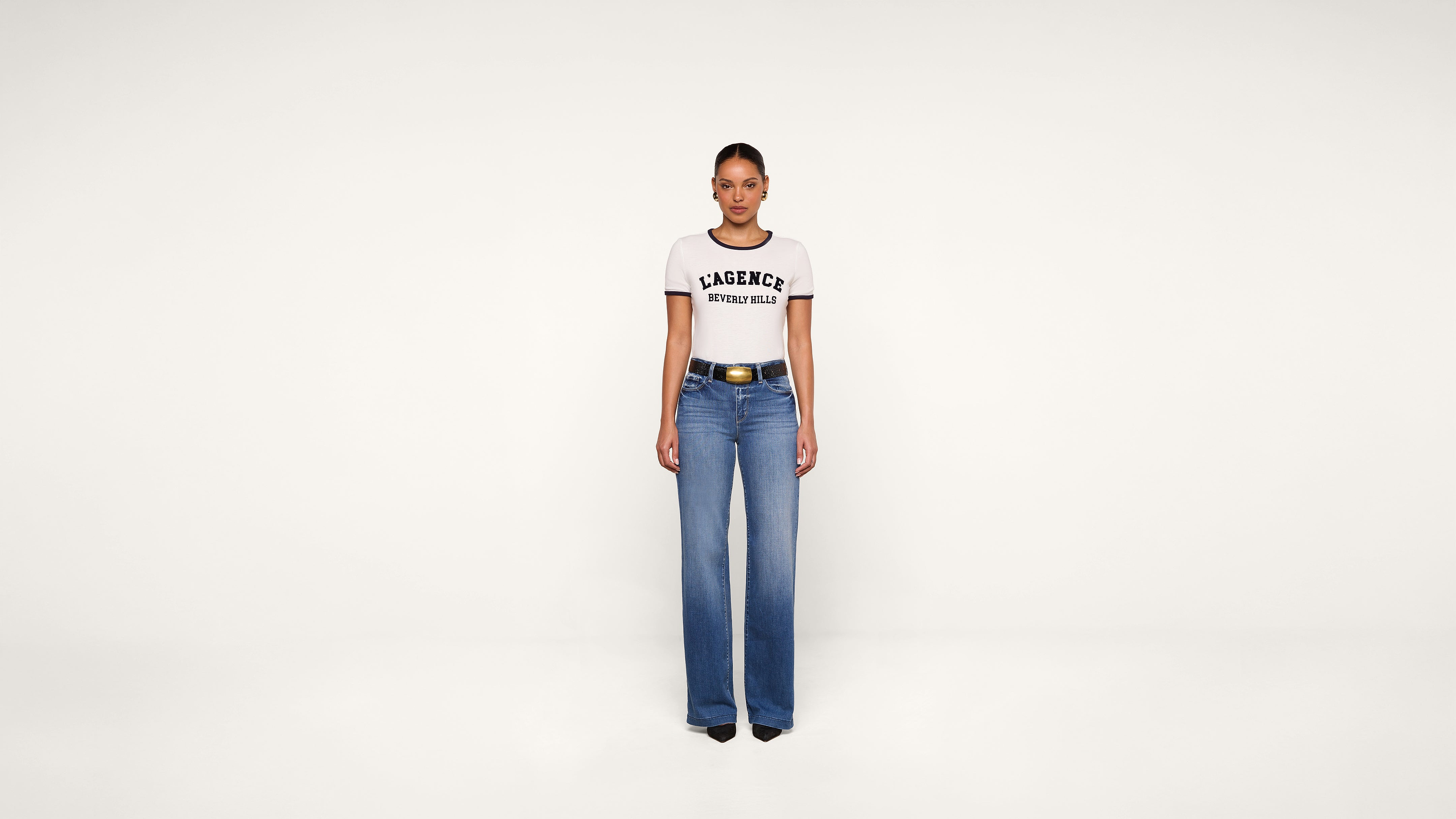 Woman stands against a plain white background, wearing a white T-shirt with black trim and LAGENCE WORLD TOUR text, blue high-waisted wide-leg jeans, a black belt with a gold buckle, and black pointed-toe shoes.
