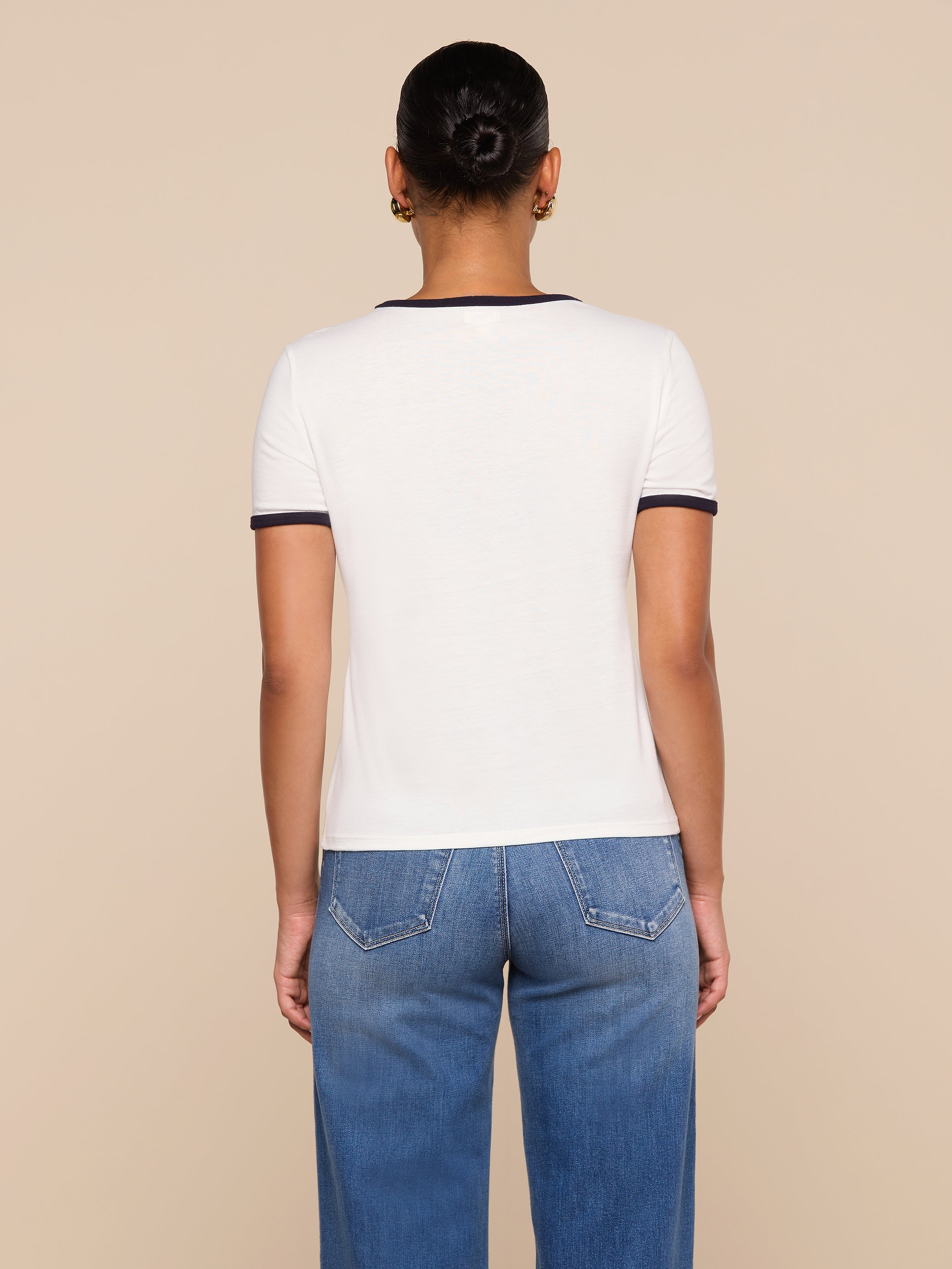 A person with short hair in a bun, wearing the white Tommie Crewneck Tee with dark trim and relaxed-fit blue jeans, stands facing away from the camera against a beige background.