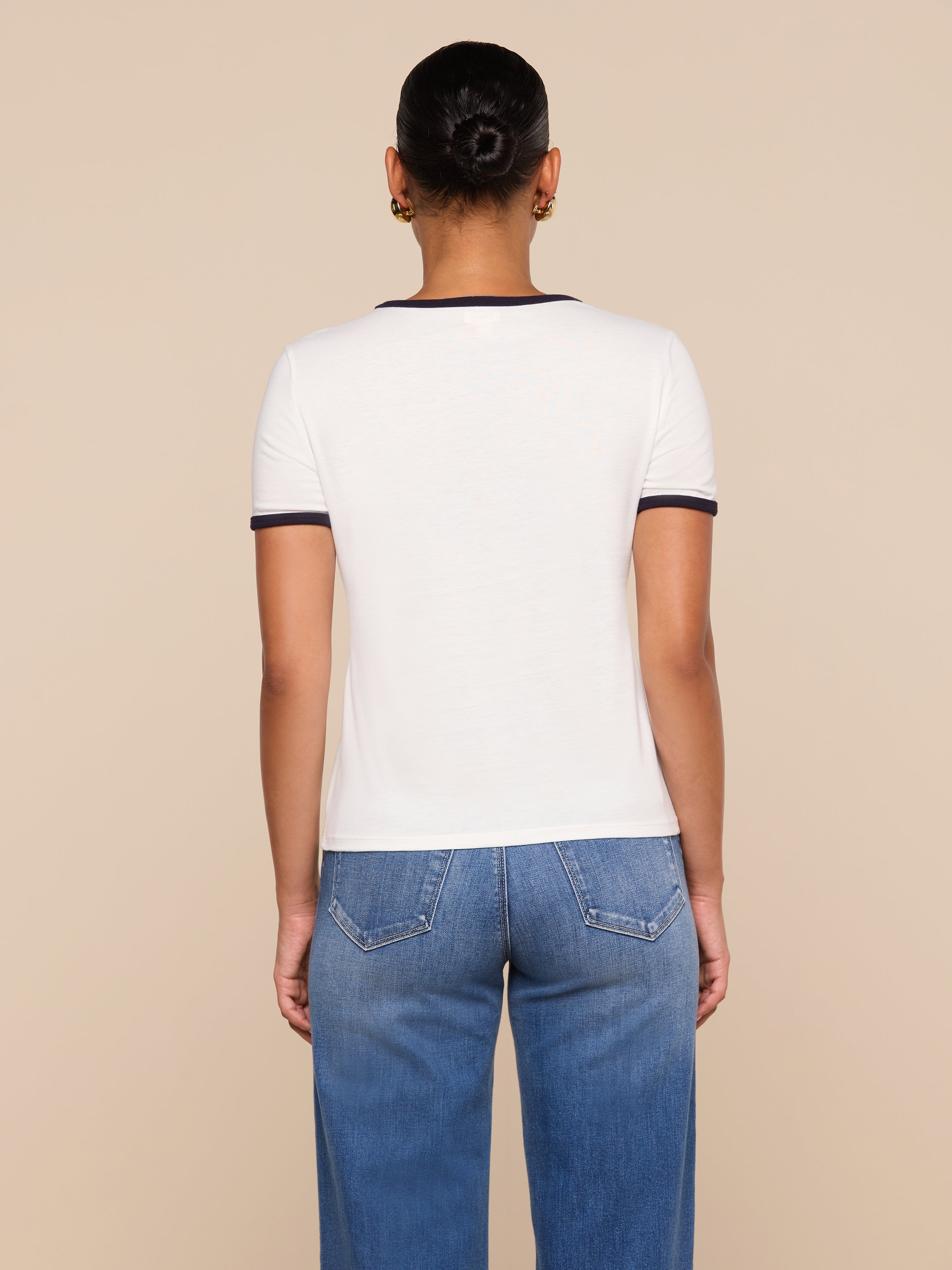 A person with short hair in a bun, wearing the white Tommie Crewneck Tee with dark trim and relaxed-fit blue jeans, stands facing away from the camera against a beige background.