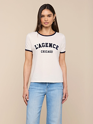 A woman stands against a plain background, wearing the Tommie Crewneck Tee—a relaxed fit white ringer tee crafted from Supima cotton with LAGENCE CHICAGO printed on it—paired with light blue jeans.