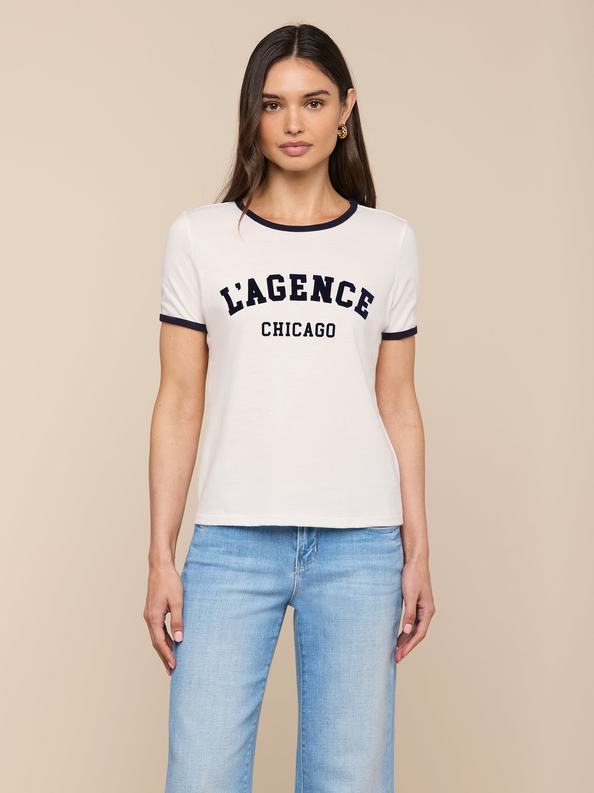 A woman stands against a plain background, wearing the Tommie Crewneck Tee—a relaxed fit white ringer tee crafted from Supima cotton with LAGENCE CHICAGO printed on it—paired with light blue jeans.