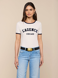 Woman with long brown hair wearing the Tommie Crewneck Tee in white, paired with light blue jeans and a black belt with a gold buckle, standing against a plain light background.