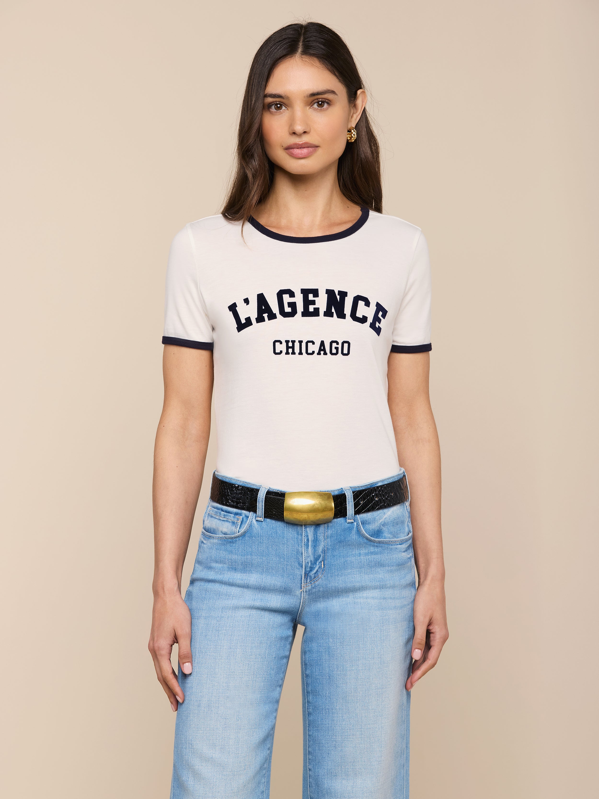 Woman with long brown hair wearing the Tommie Crewneck Tee in white, paired with light blue jeans and a black belt with a gold buckle, standing against a plain light background.