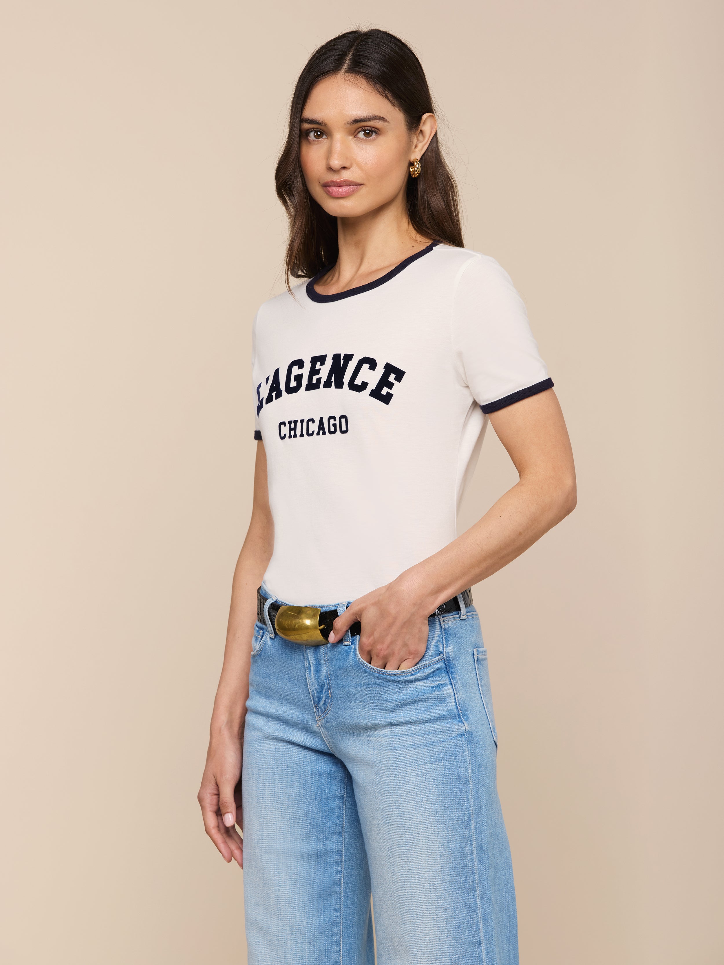 A woman with long brown hair wears the Tommie Crewneck Tee—a relaxed fit white ringer shirt with “AGENCE CHICAGO” on the front—paired with light blue jeans and a black belt, posing with one hand in her pocket against a plain background.