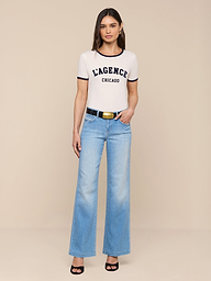 A woman stands against a plain white background wearing a white Tommie Crewneck Tee made from Supima cotton, light blue wide-leg jeans, a black belt, and black high-heeled sandals. She faces forward with her arms relaxed at her sides.