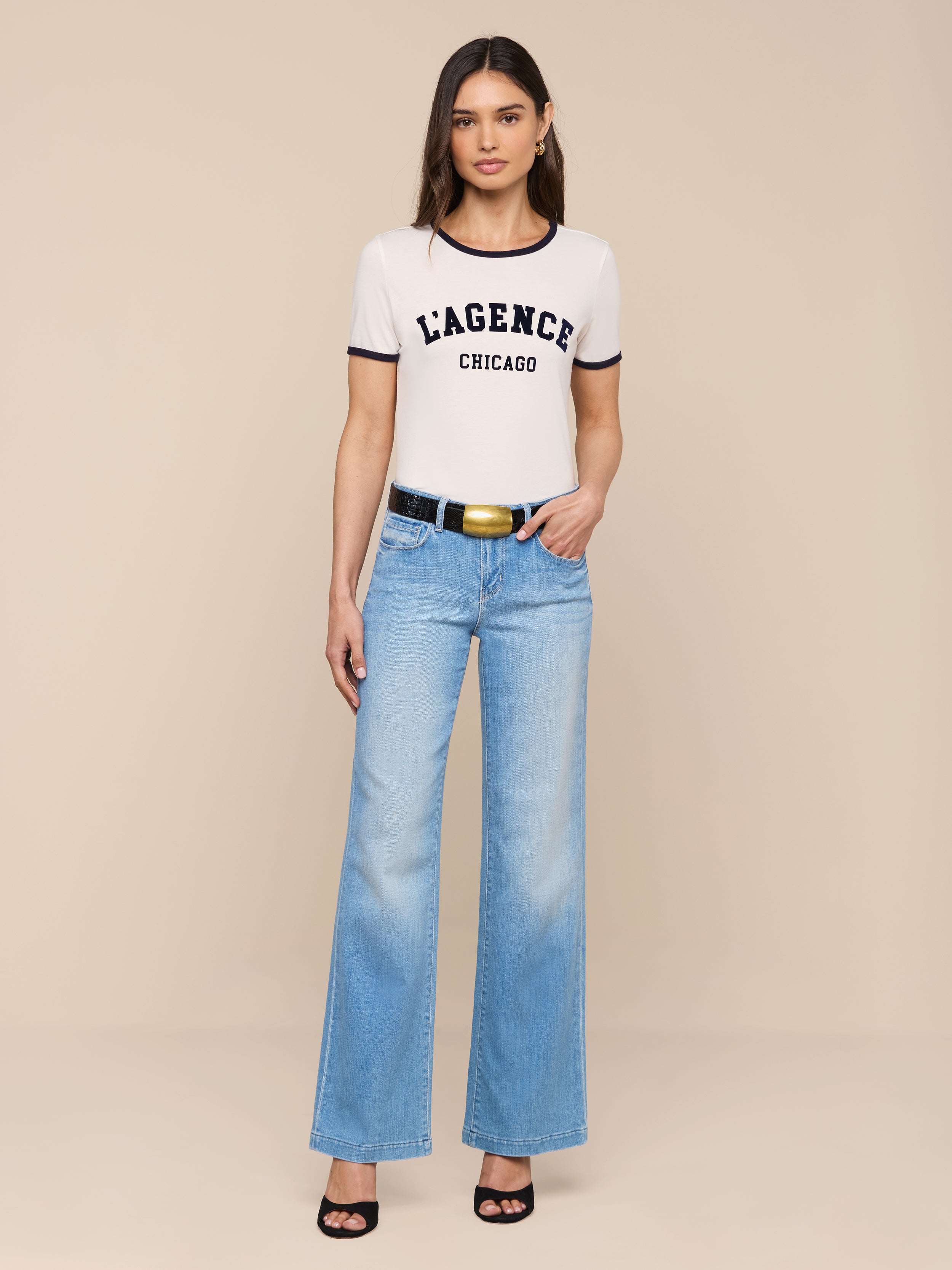 A woman stands against a plain white background wearing a white Tommie Crewneck Tee made from Supima cotton, light blue wide-leg jeans, a black belt, and black high-heeled sandals. She faces forward with her arms relaxed at her sides.