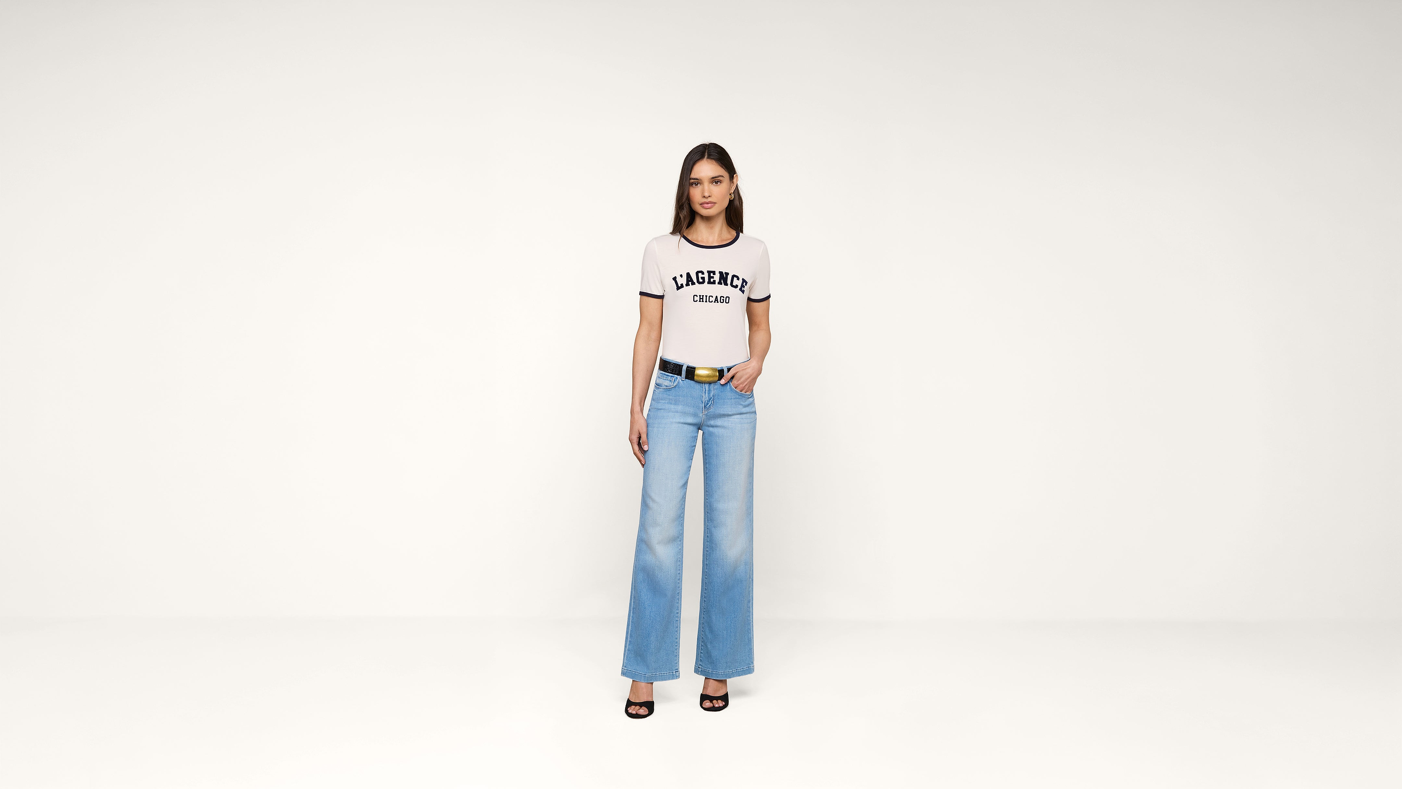 A woman stands against a plain white background, wearing a white LAGERFELD t-shirt, light blue wide-leg jeans, black belt, and black open-toe heels. She faces forward with her hands relaxed by her sides.