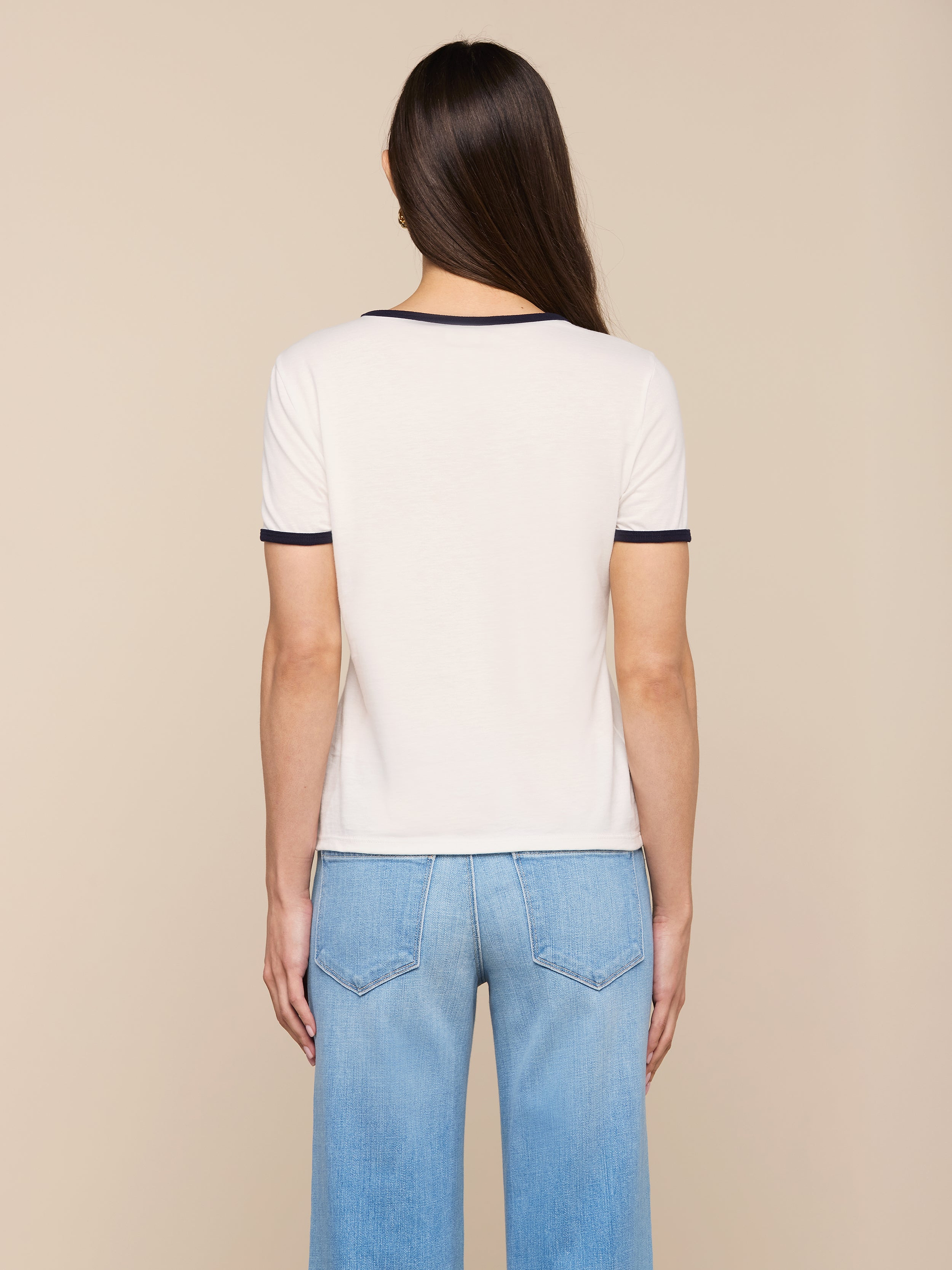 A woman with long brown hair stands facing away, wearing the Tommie Crewneck Tee— a white ringer style with dark trim—paired with light blue jeans against a plain light background.