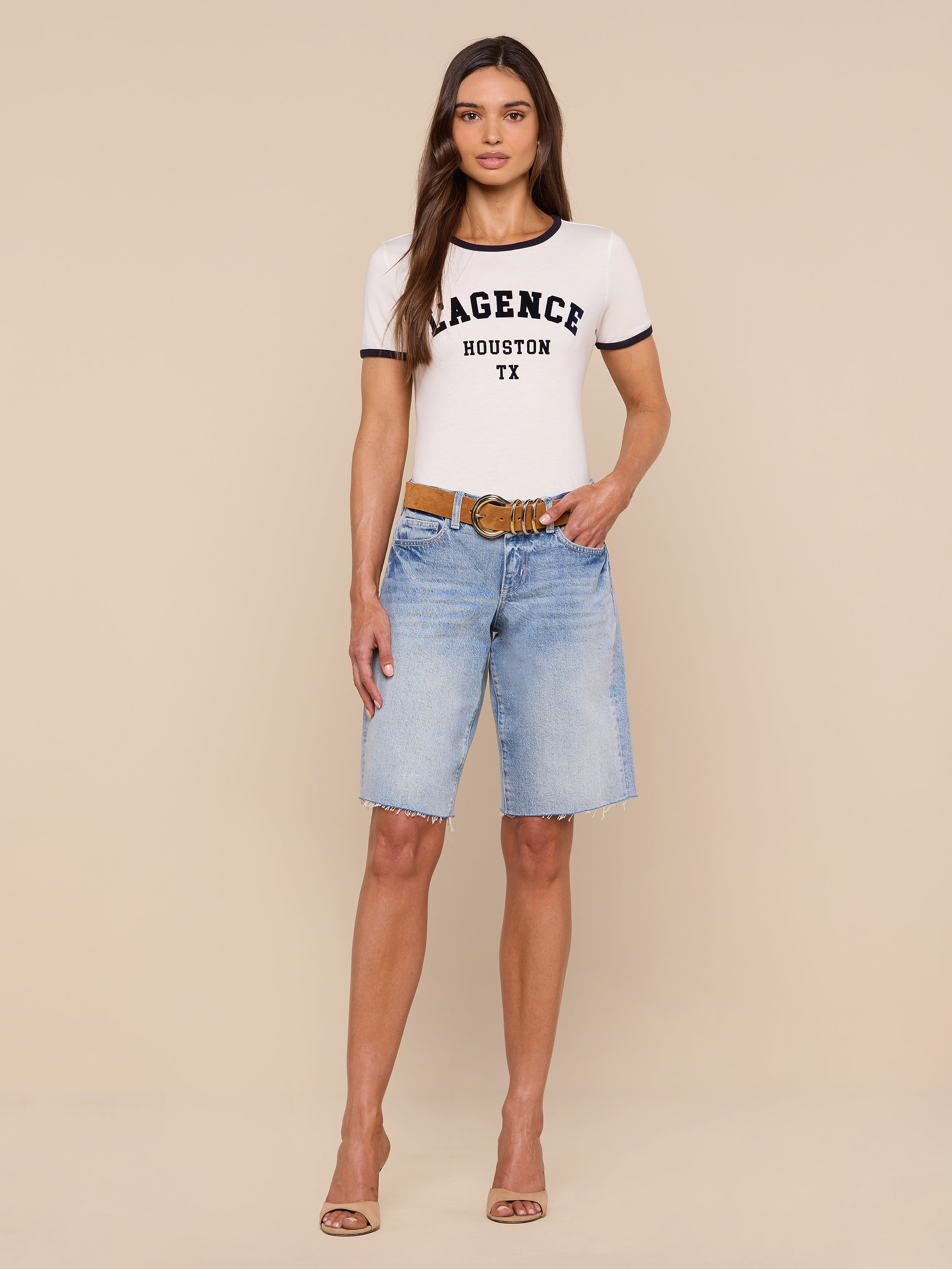 A woman stands facing forward, wearing a relaxed fit Tommie Crewneck Tee—a white T-shirt with black trim and AGENCE HOUSTON TX text—paired with light blue knee-length denim shorts, a brown belt, and beige heels. The background is plain white.