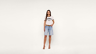 A woman stands against a plain white background, wearing a white graphic T-shirt that says JAGGER DENIM TX, light blue knee-length denim shorts, a brown belt, and tan high-heeled sandals.