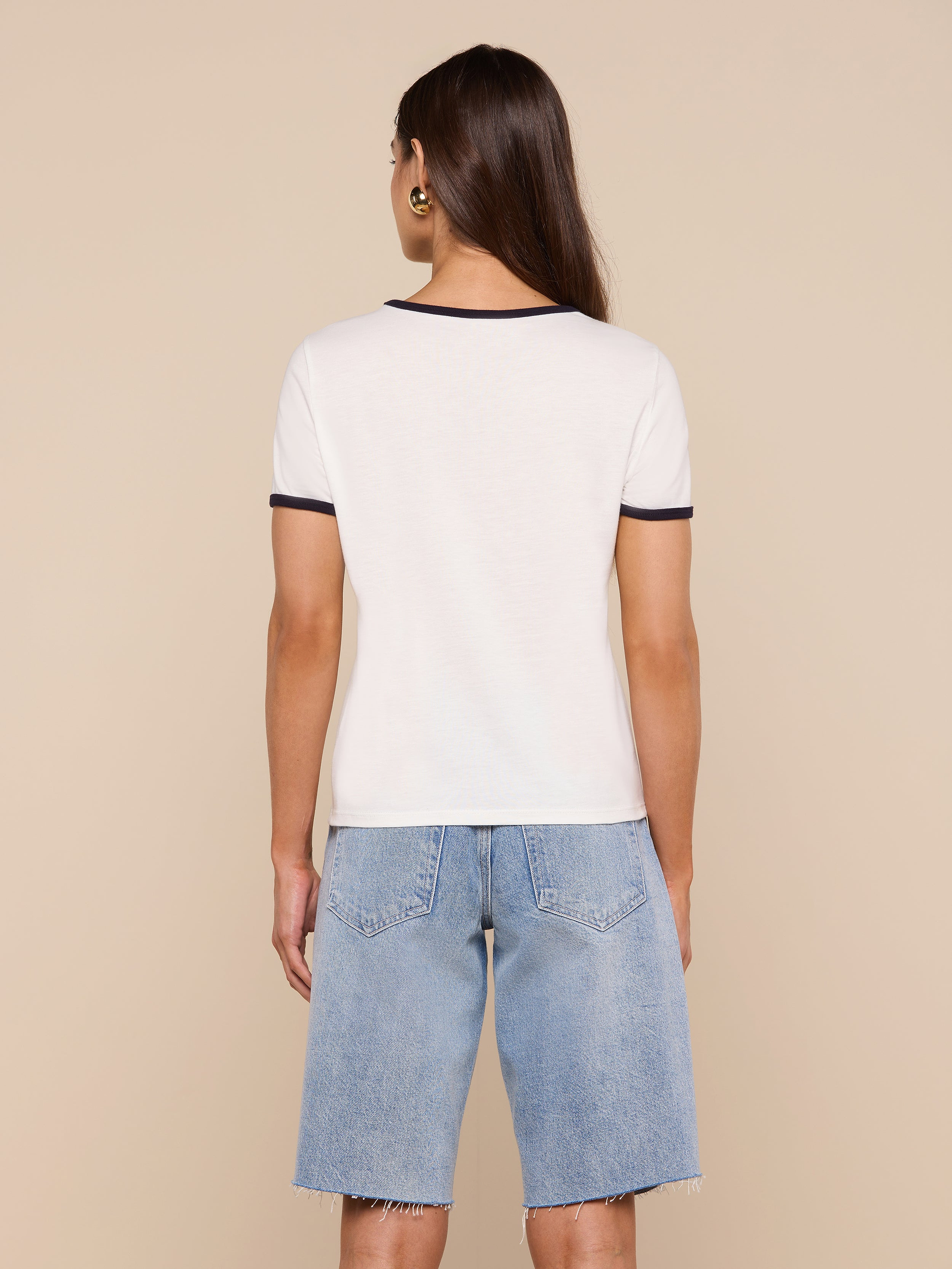 Wearing the relaxed fit Tommie Crewneck Tee in white with dark trim, a person faces away against a plain light background, paired with light blue knee-length denim shorts.