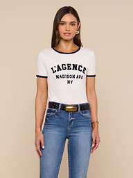 A woman with long brown hair wears a white Tommie Crewneck Tee made from Supima cotton, blue jeans, a black belt with a gold buckle, and gold earrings, standing against a beige background.