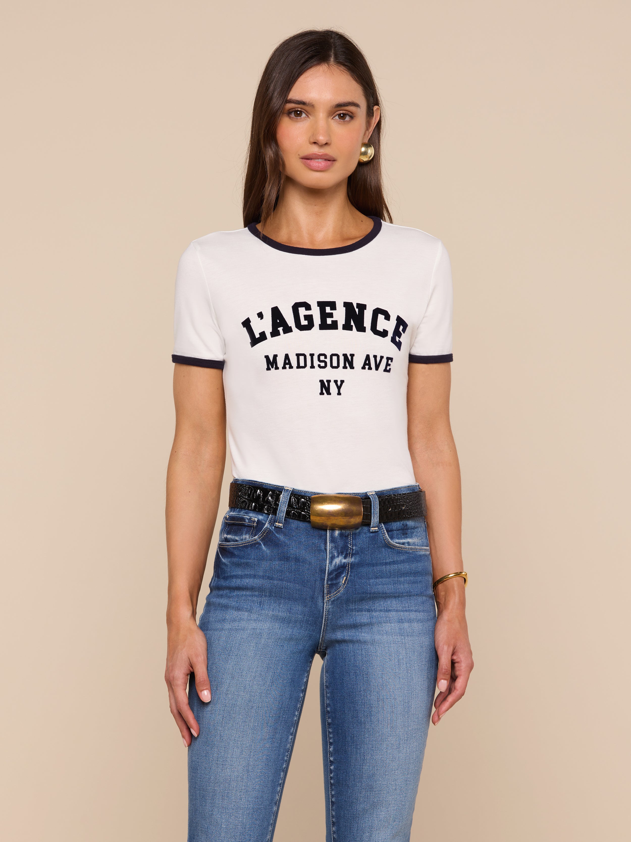 A woman with long brown hair wears a white Tommie Crewneck Tee made from Supima cotton, blue jeans, a black belt with a gold buckle, and gold earrings, standing against a beige background.