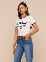 A woman with long brown hair wears a relaxed fit Tommie Crewneck Tee made of Supima cotton, blue jeans, and gold accessories, standing against a beige background.