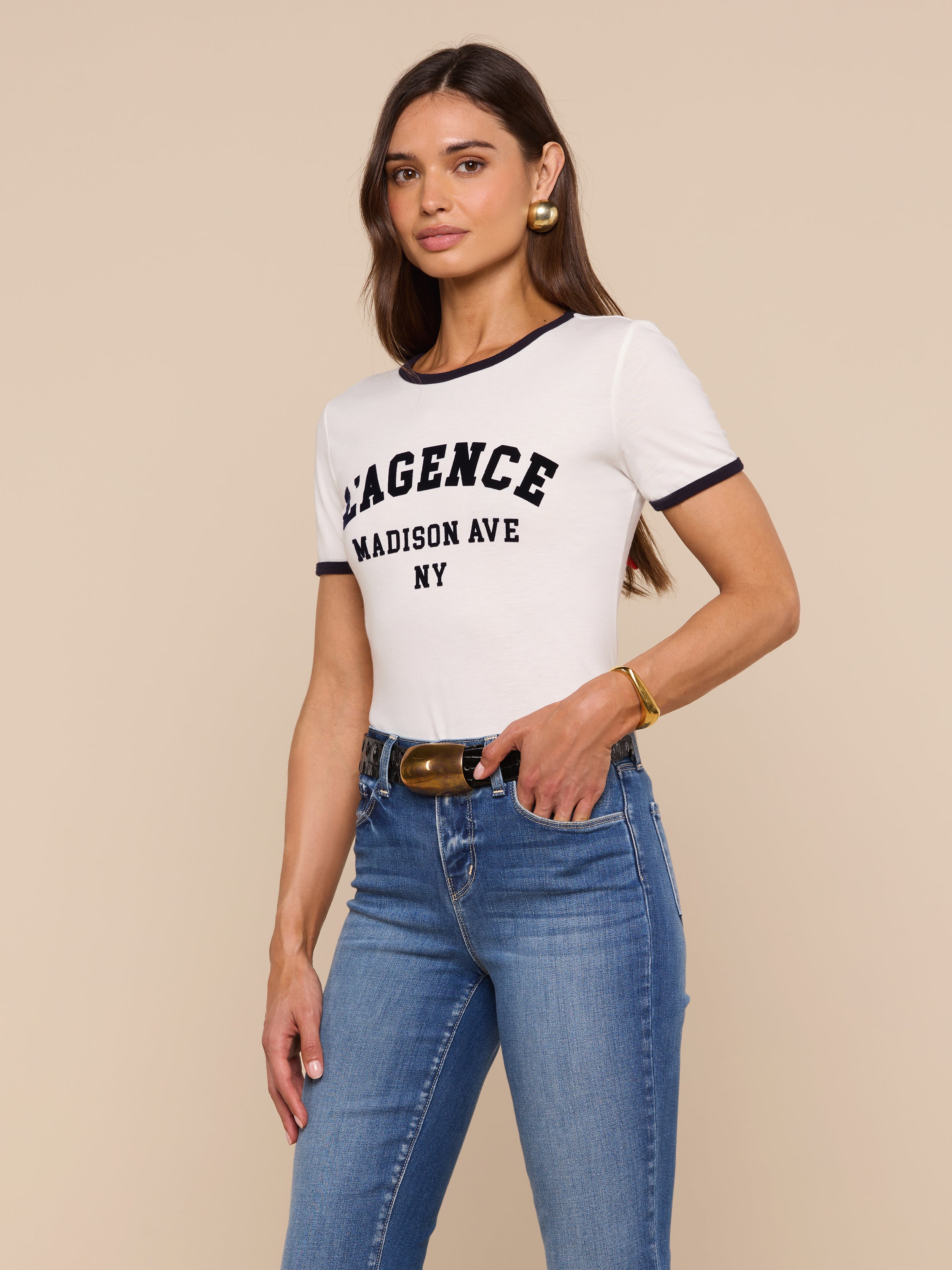 A woman with long brown hair wears a relaxed fit Tommie Crewneck Tee made of Supima cotton, blue jeans, and gold accessories, standing against a beige background.