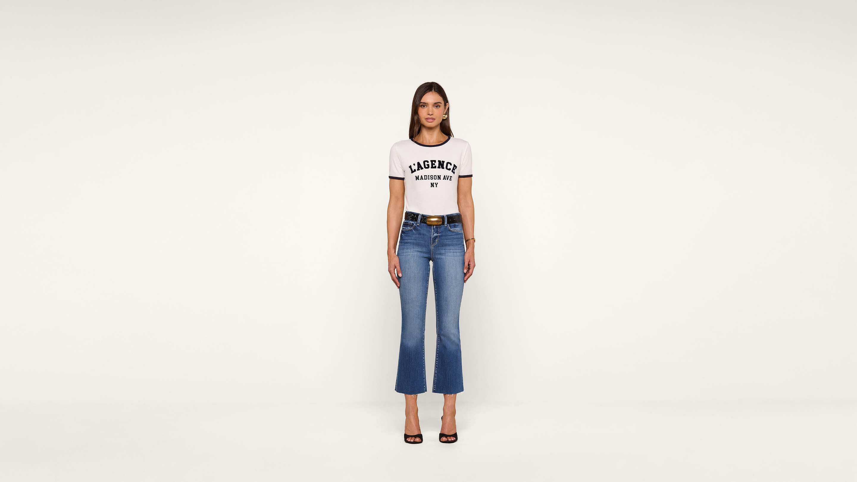 A woman stands in front of a plain white background wearing a white graphic t-shirt, blue cropped flare jeans, a black belt, and black heeled sandals. She faces forward with her arms at her sides.