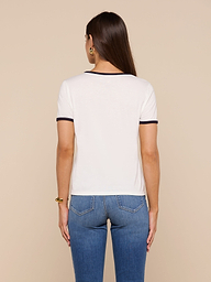 A woman with long brown hair stands with her back to the camera, wearing a relaxed fit Tommie Crewneck Tee in white with black trim and blue jeans against a beige background.