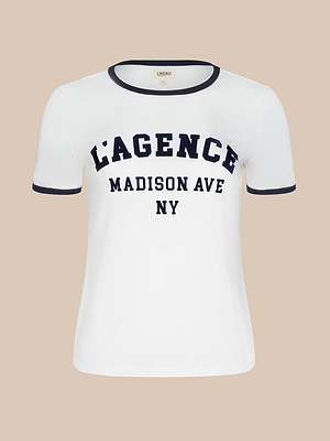 The Tommie Crewneck Tee is a white short-sleeve shirt with black trim on the collar and sleeves, crafted from Supima cotton. It features bold L’AGENCE MADISON AVE NY text on the front and is shown against a beige background.