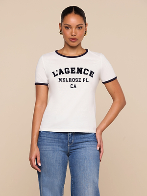 A woman wearing the Tommie Crewneck Tee, a relaxed fit Supima cotton shirt with LAGENCE MELROSE PL CA text, and blue jeans stands against a plain beige background.