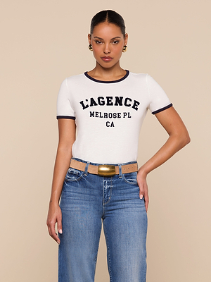 A woman stands against a beige background wearing the Tommie Crewneck Tee—a relaxed fit white Supima cotton ringer with LAGENCE MELROSE PL CA in black letters—paired with blue jeans, a tan belt with a gold buckle, and gold earrings. Hand on her hip.