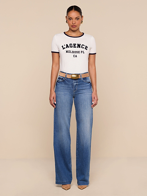 A person stands facing forward, wearing a white Tommie Crewneck Tee with a relaxed fit, blue high-waisted wide-leg jeans, a brown belt, tan pointed shoes, and gold earrings against a plain beige background.