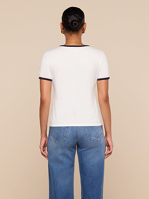 A person stands facing away in a white Tommie Crewneck Tee with dark trim and blue jeans. Their hair is styled in a neat bun. The relaxed-fit shirt contrasts with the plain beige background.