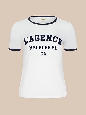 The Tommie Crewneck Tee is a white ringer shirt with black trim on the collar and sleeves, featuring bold LAGENCE MELROSE PL CA text on the front. Made from soft Supima cotton for a relaxed fit and all-day comfort.